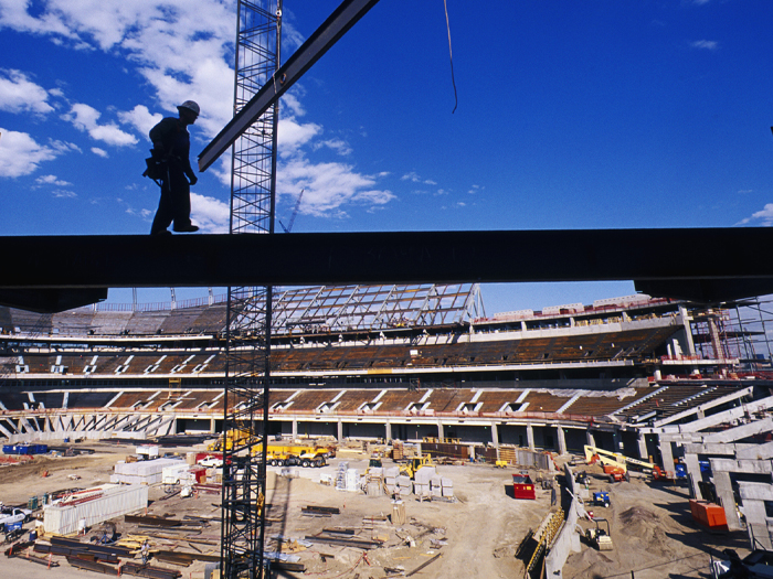 A worker walks along a girder suspended above a stadium construction site