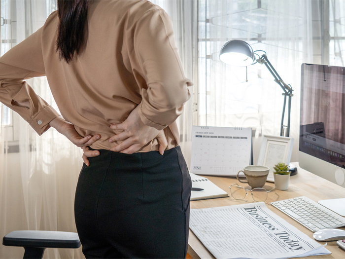 A woman stands in front of her desk, holding her back to indicate back strain