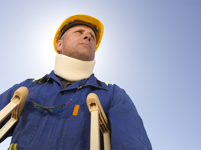 A worker in coveralls and a hardhat stands on crutches